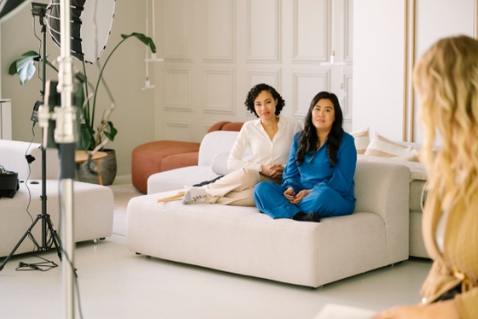 Twee vrouwen zitten op een lichte bank in een modern ingerichte studio terwijl ze worden gefilmd of geïnterviewd.