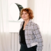 Vrouw met rossig krullend haar poseert binnen voor een wit gordijn, gekleed in een geruite blazer.