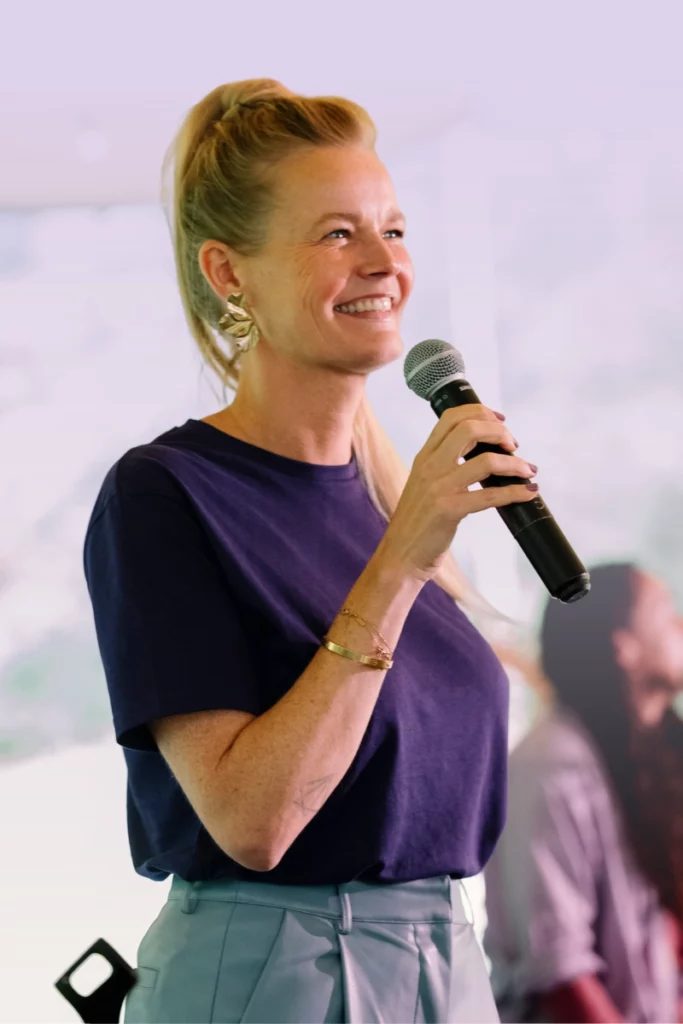 Vrouw spreekt glimlachend in een microfoon op een podium tijdens een presentatie of evenement.