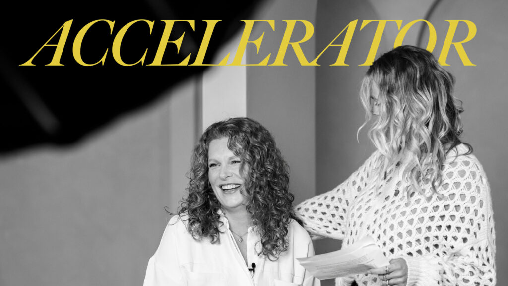 Twee vrouwen lachen en praten tijdens een sessie van het leiderschap vrouwen programma, met het woord "ACCELERATOR" groot in beeld.