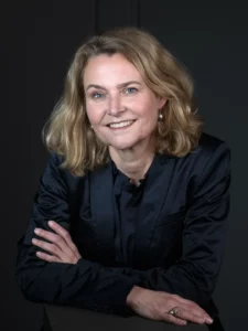 Portret van Margreet, een vrouw met blond haar in een donkerblauwe blouse, die glimlachend en met gekruiste armen voor een donkere achtergrond poseert.