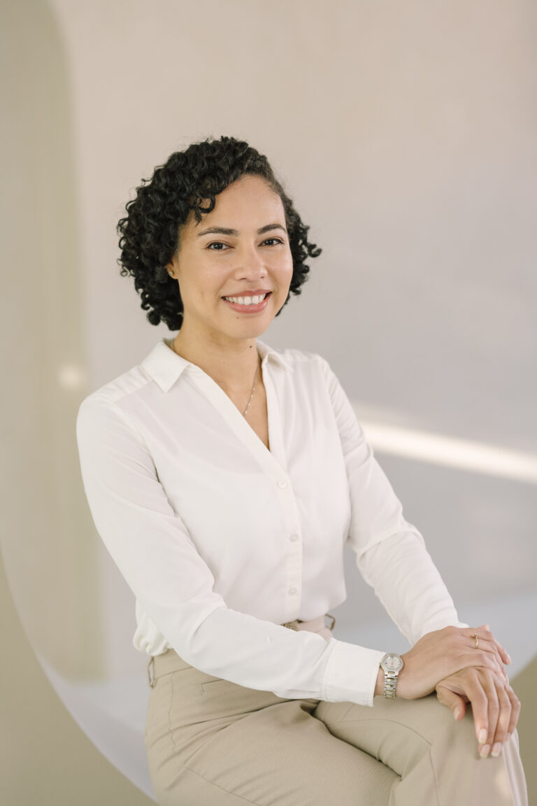 Vrouw met krullend haar zit zelfverzekerd op een stoel in witte blouse en beige broek.