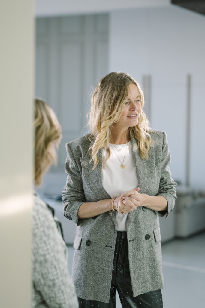 Vrouwelijke leider in een grijze blazer spreekt met een collega in een moderne kantoorruimte.