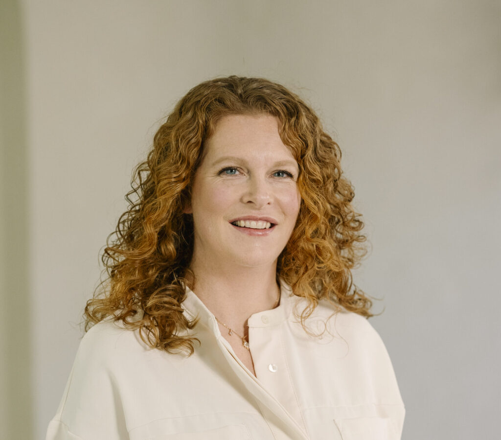 Portret van een glimlachende vrouw met rood krullend haar in een lichte blouse, tegen een neutrale achtergrond.