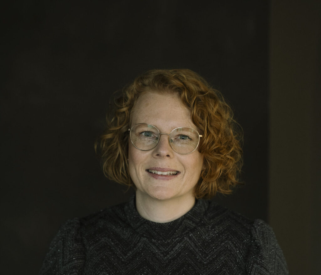 Portret van een vrouw met rood krullend haar en een bril, tegen een donkere achtergrond.