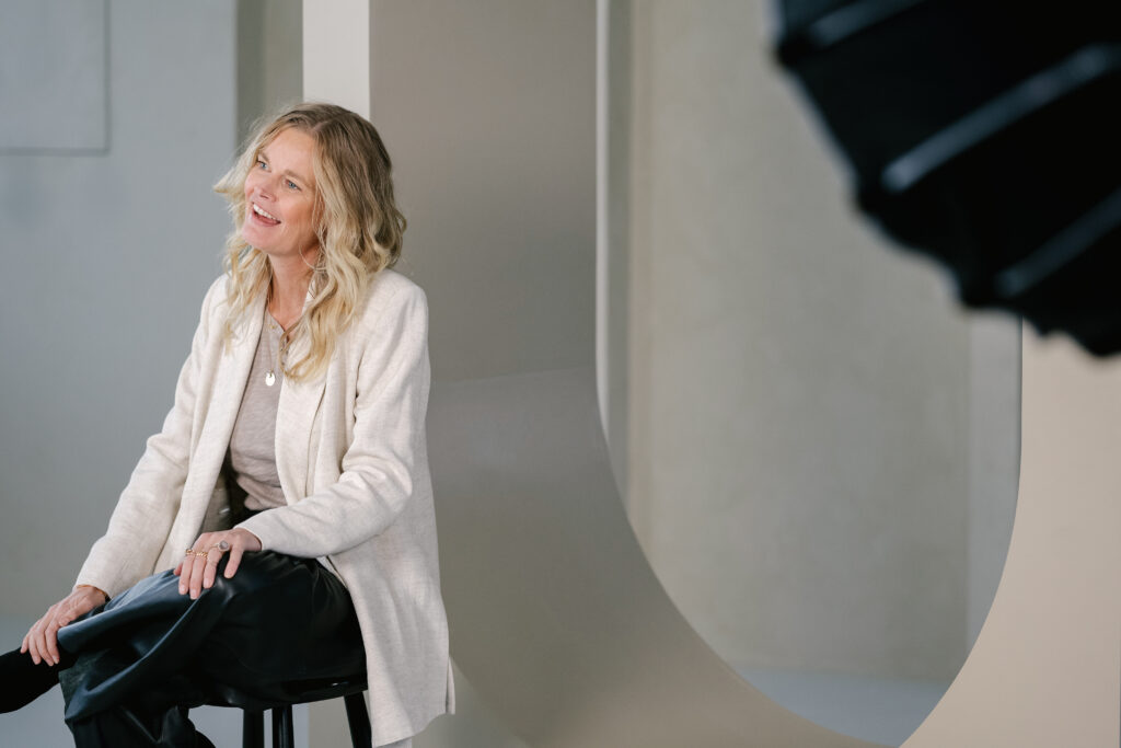 Vrouw met blond haar zit glimlachend en ontspannen op een kruk in een moderne studio-opstelling.