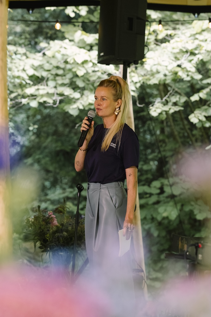 Vrouw spreekt op een podium in een groene, natuurlijke omgeving met microfoon in de hand.