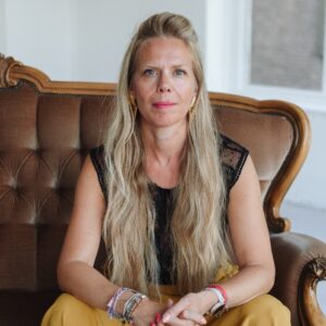 Vrouwelijke spreker met lange blonde haren zit op een vintage bank en kijkt recht in de camera.