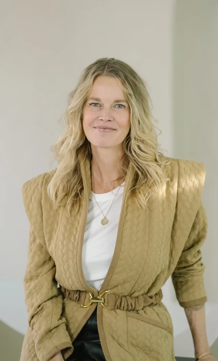Vrouw met blond, golvend haar draagt een beige gewatteerde blazer en witte top, en glimlacht rustig naar de camera.