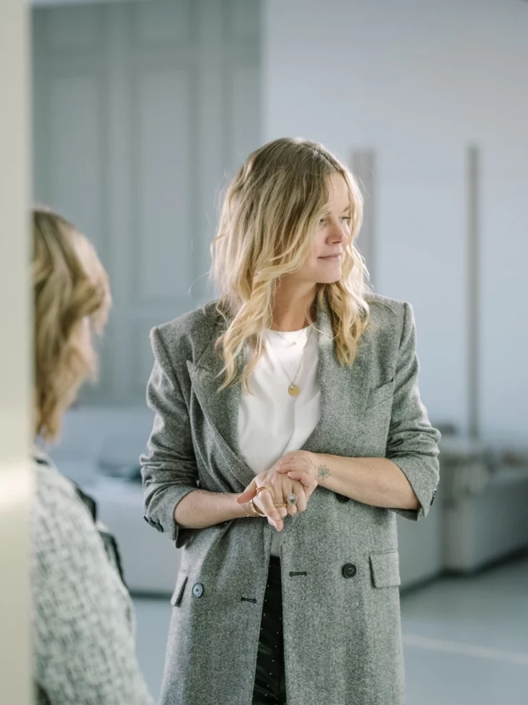 Vrouw in grijze blazer staat in een moderne ruimte en kijkt opzij tijdens een gesprek.