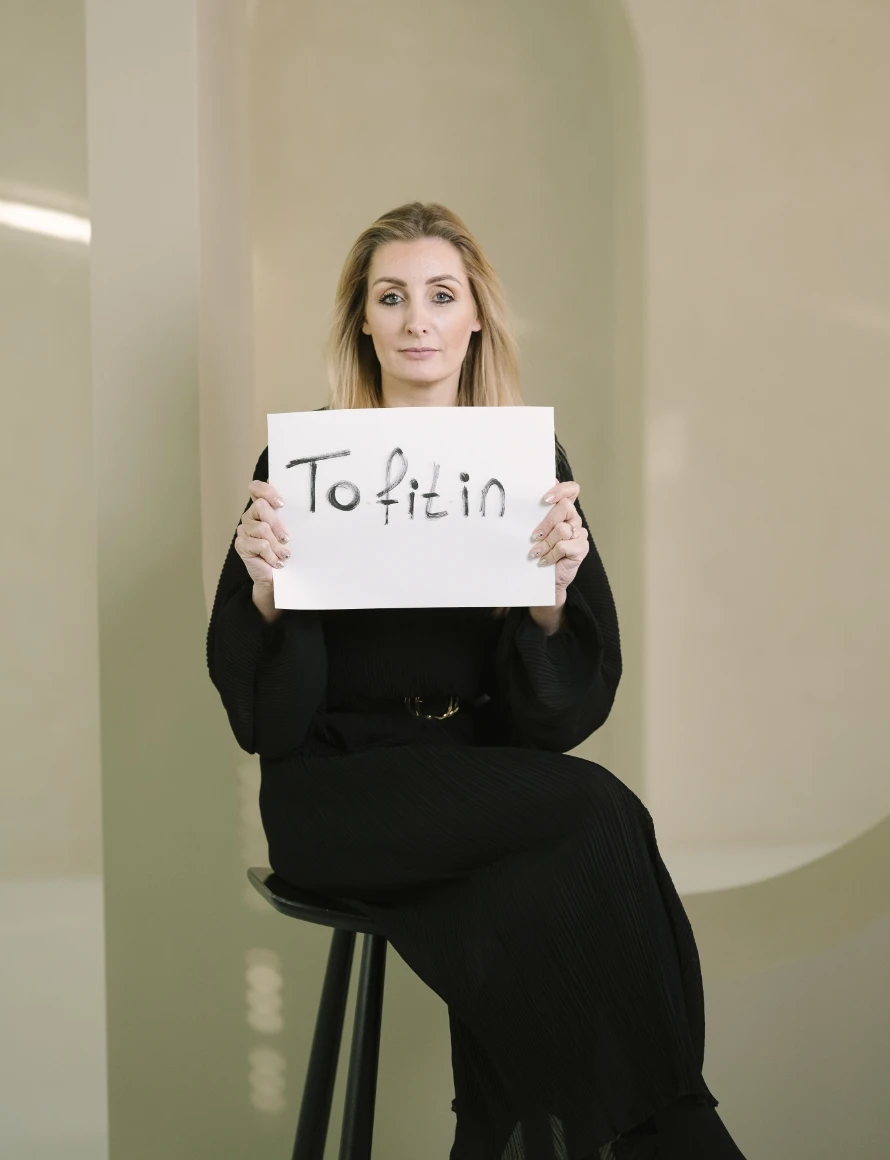 Vrouw in zwarte kleding houdt een bord vast met de handgeschreven tekst "To fit in".