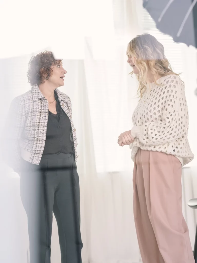 Twee vrouwen in gesprek tijdens een fotoshoot voor The Accelerator, met zachte belichting en een witte achtergrond.