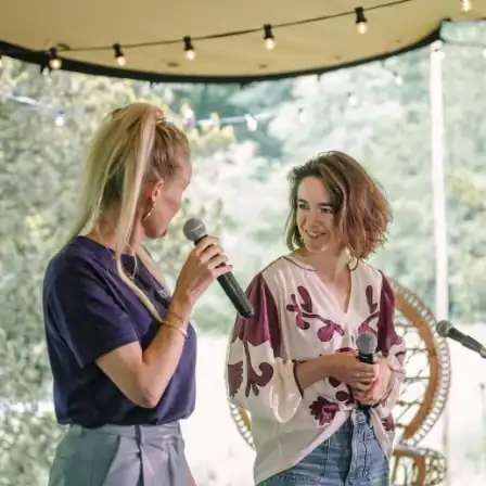 Twee vrouwen spreken op een buitenpodium tijdens het New Female Leaders Festival, met microfoons in de hand.