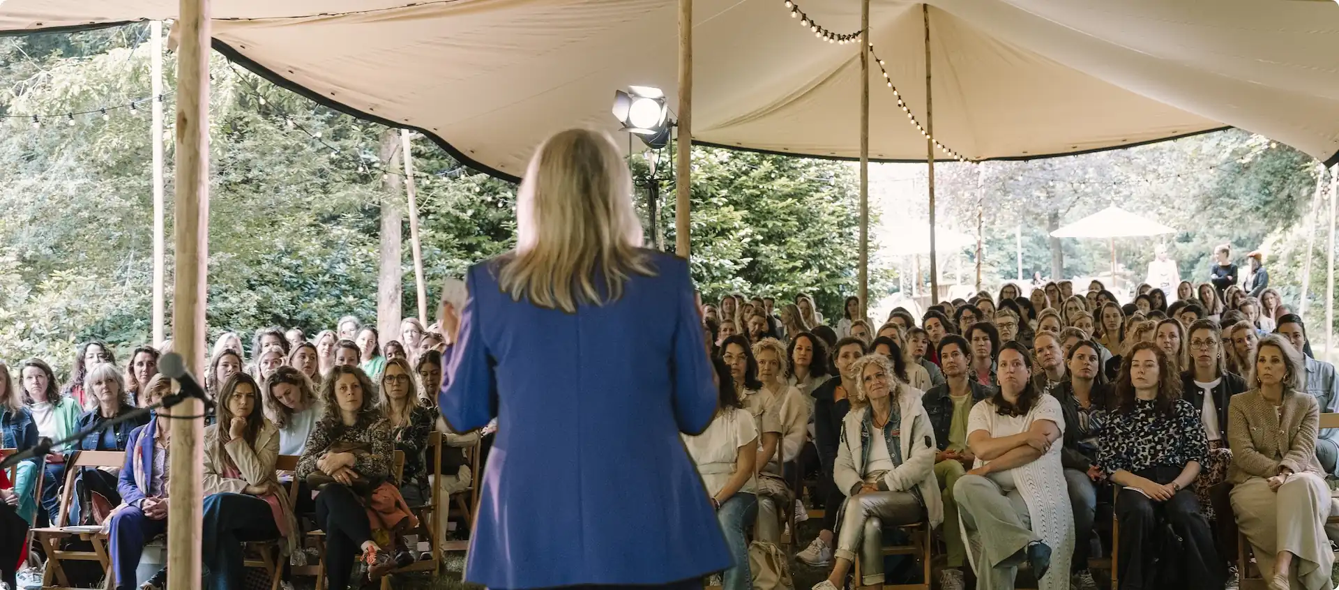 Vrouw spreekt voor een aandachtig publiek tijdens het New Female Leaders Festival onder een tent in een groene buitenomgeving.