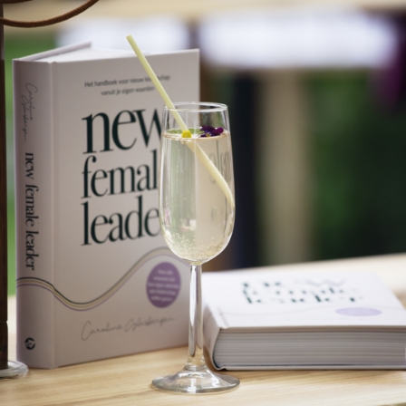Een glas bruisende drank met een rietje en bloemetje staat op tafel naast het boek "New Female Leader".