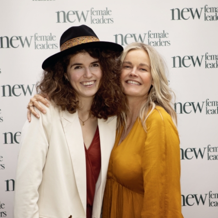 Twee vrouwen poseren lachend voor een fotowand met het logo van "New Female Leaders" tijdens een evenement.